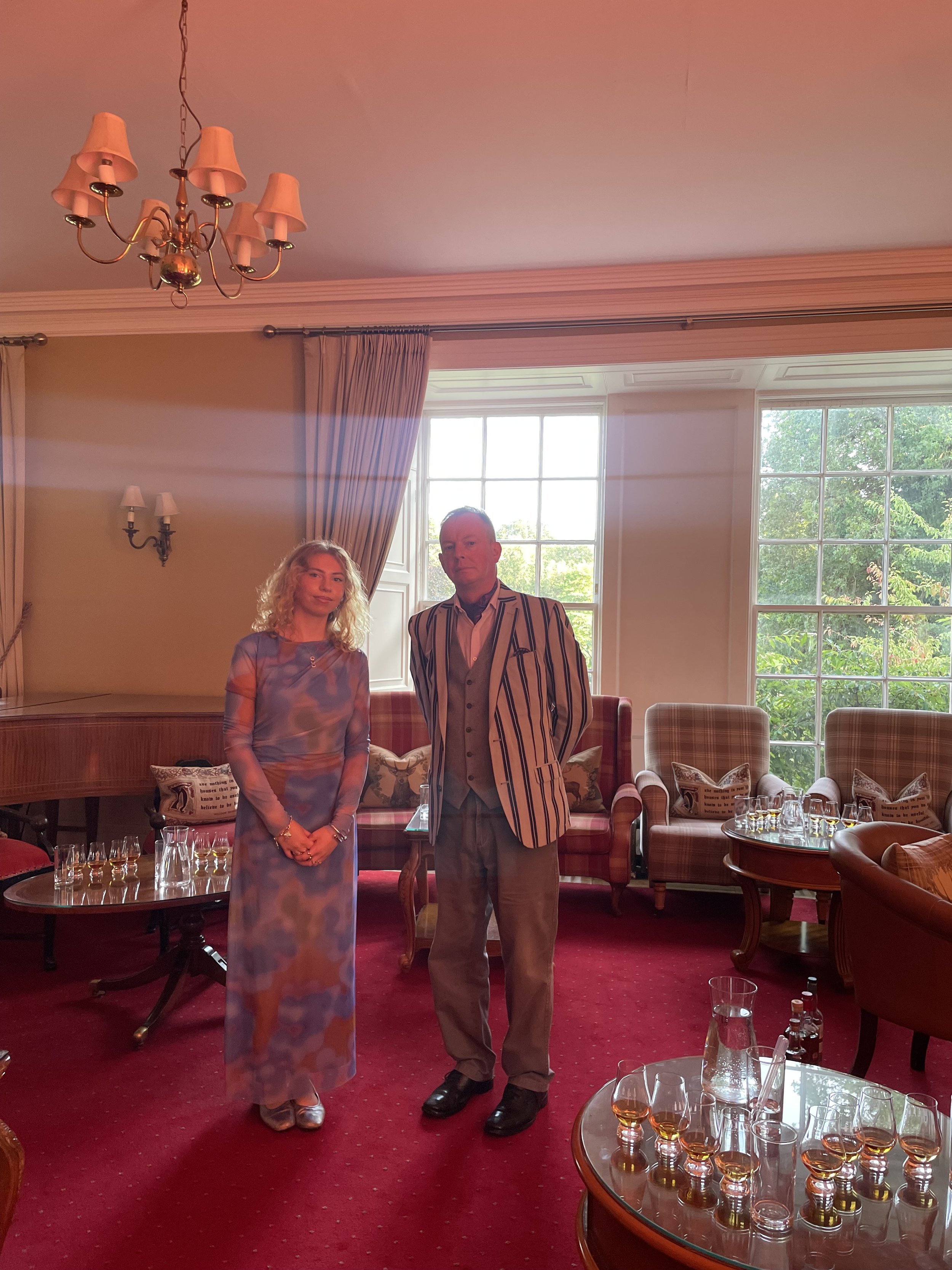 Two people standing in a well-lit room with large windows, red carpet, and classic furniture, decorated for a gathering or celebration.