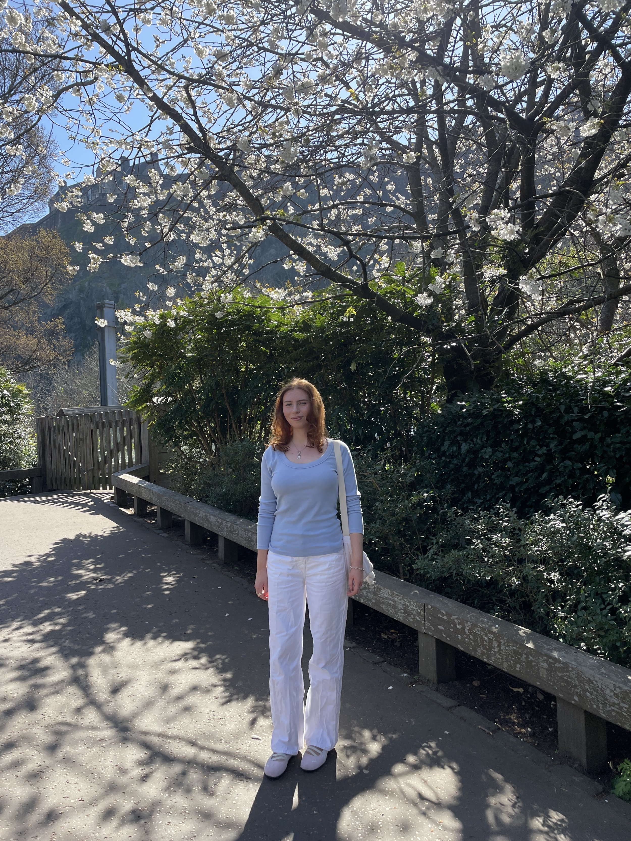 A woman stands outdoors on a sunny day with blooming tree branches overhead and greenery behind her, wearing a light blue top and white pants.