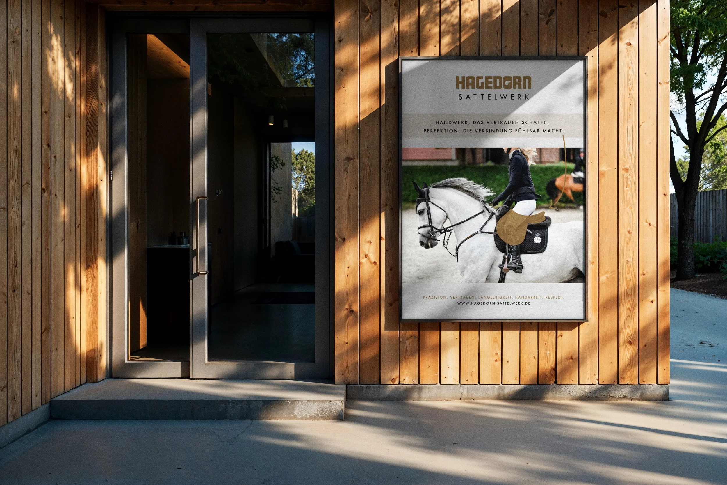Wood-paneled exterior entrance to a building with a glass door on the left. On the right, a large poster advertising Hagedorn Sattelwerk features a person riding a white horse during daylight, with trees and a garden in the background.