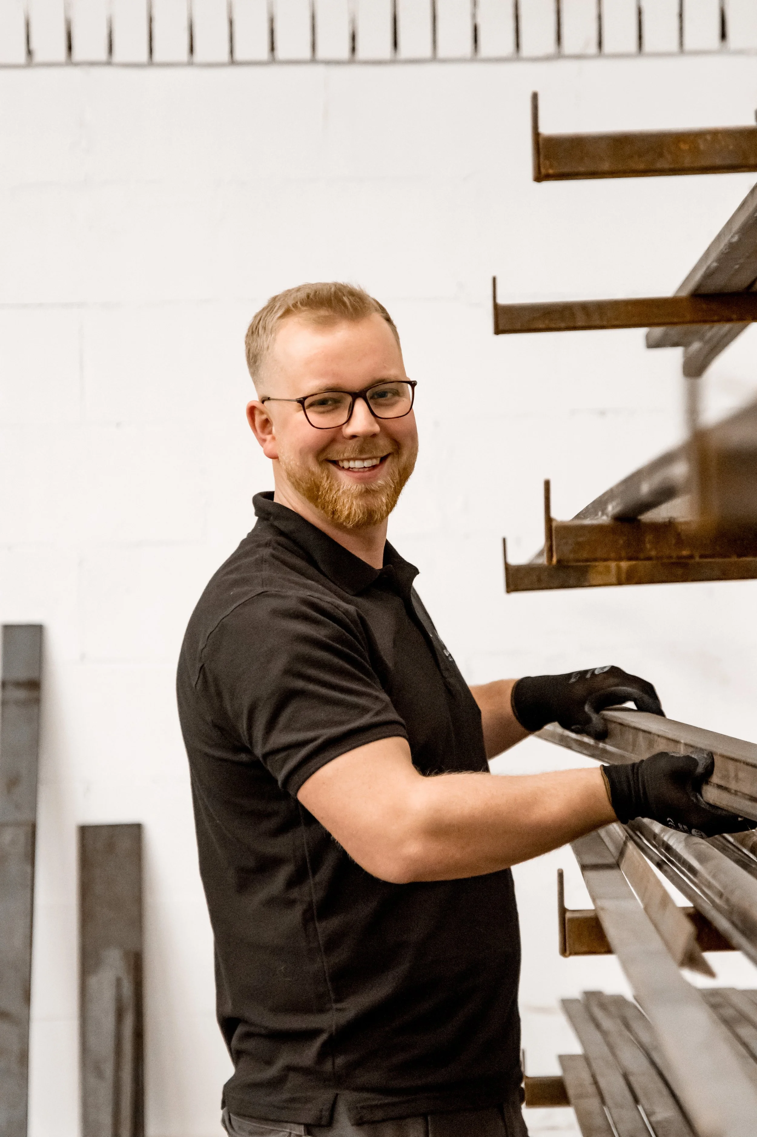 A young man with glasses and a beard smiling while working with metal on a shelf in a workshop.