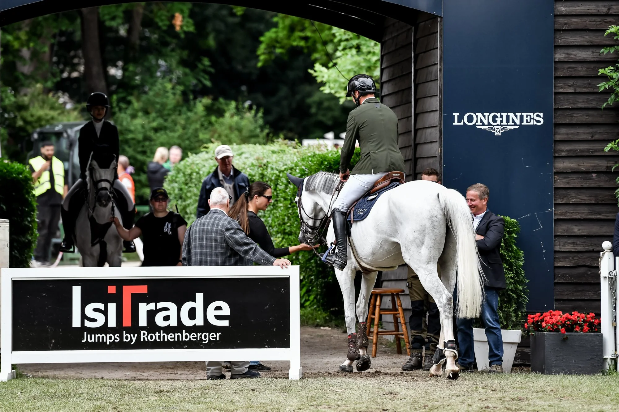 A man in riding attire mounted on a white horse, shaking hands with a woman, as others gather around at a horse jumping event marked by a sign that reads 'IsiTrade Jumps by Rothenberger,' with trees and spectators in the background.
