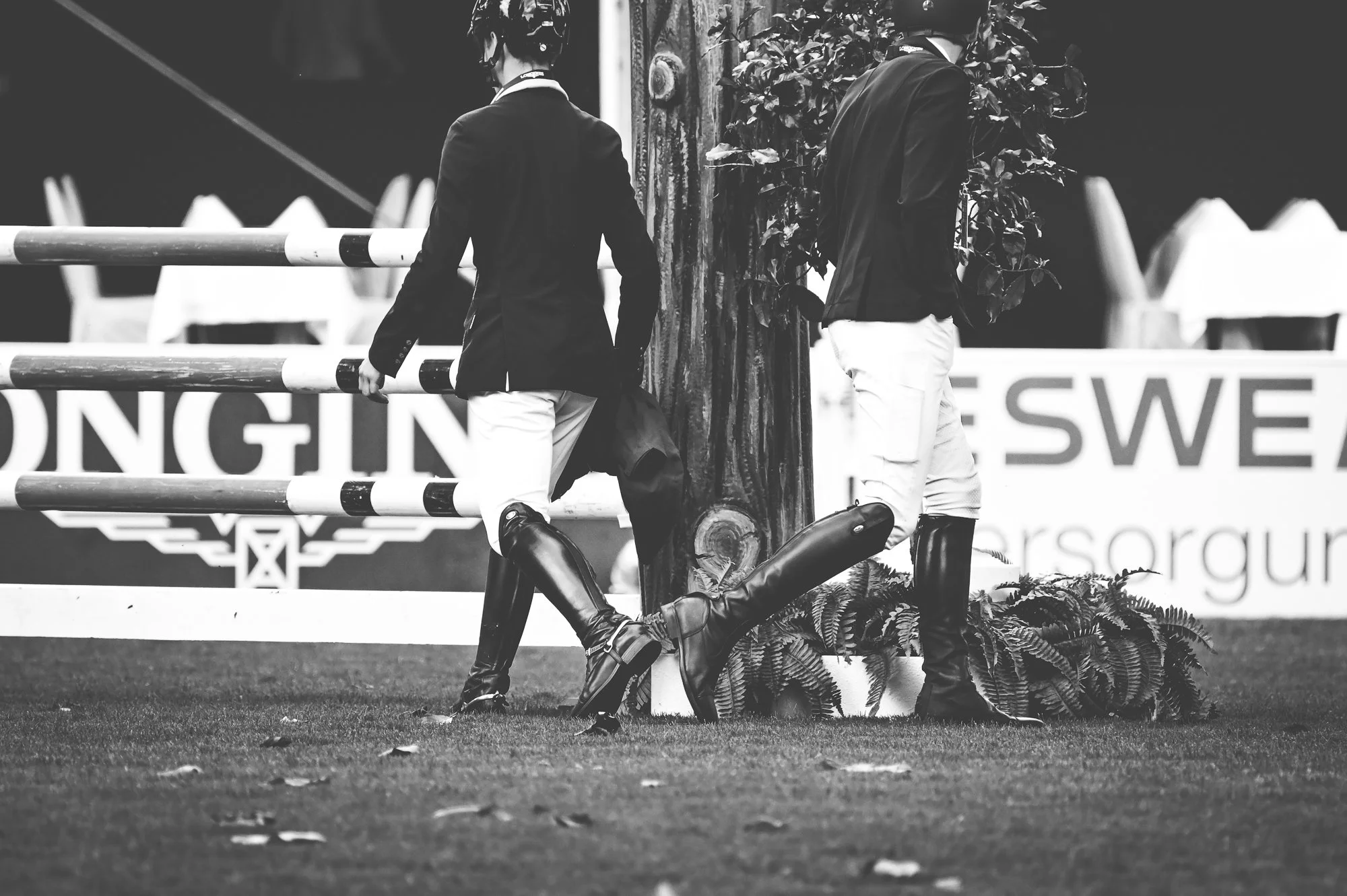 Two equestrians in riding gear stand next to a tree on a grassy field, with one leaning against the tree and the other holding the reins, in an outdoor arena.