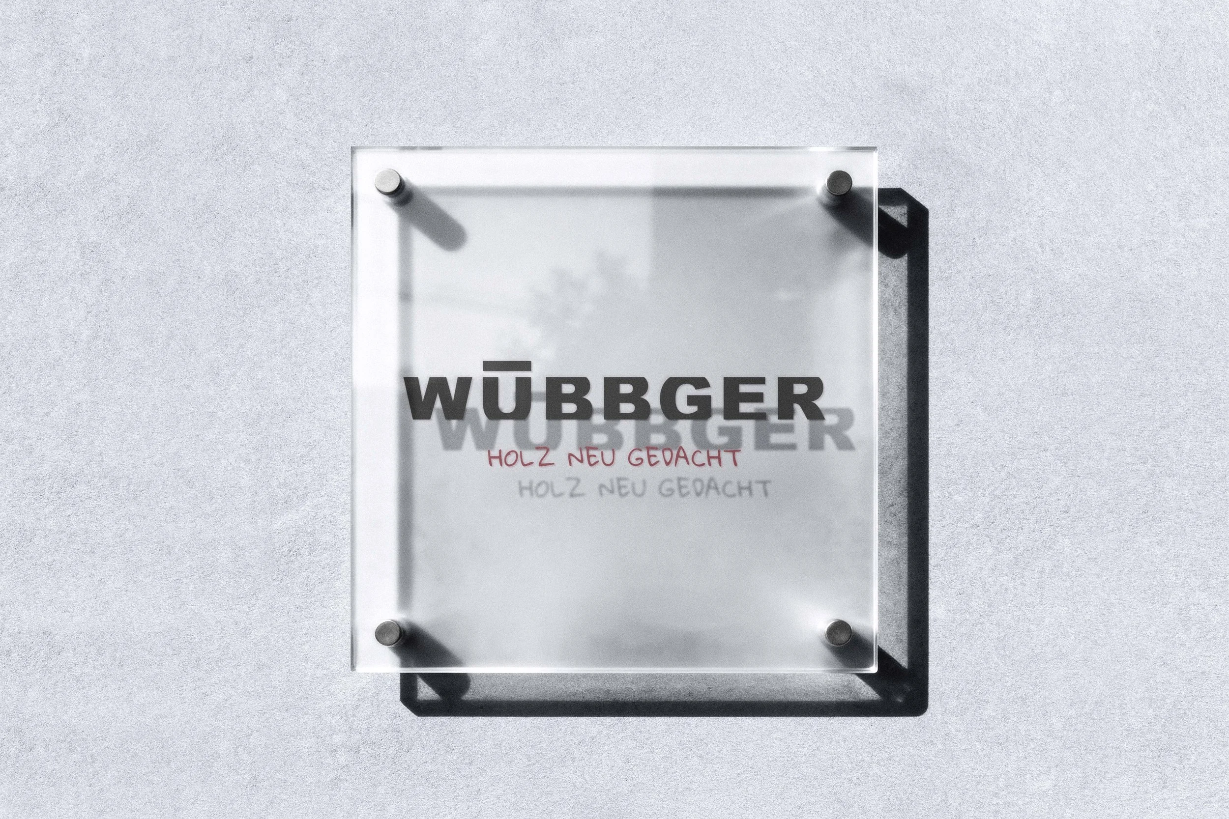 A glass sign on a wall with the word WÜBBGER, and the phrase HOLZ NEU GEDACHT below it, casting shadows from the mounting bolts.