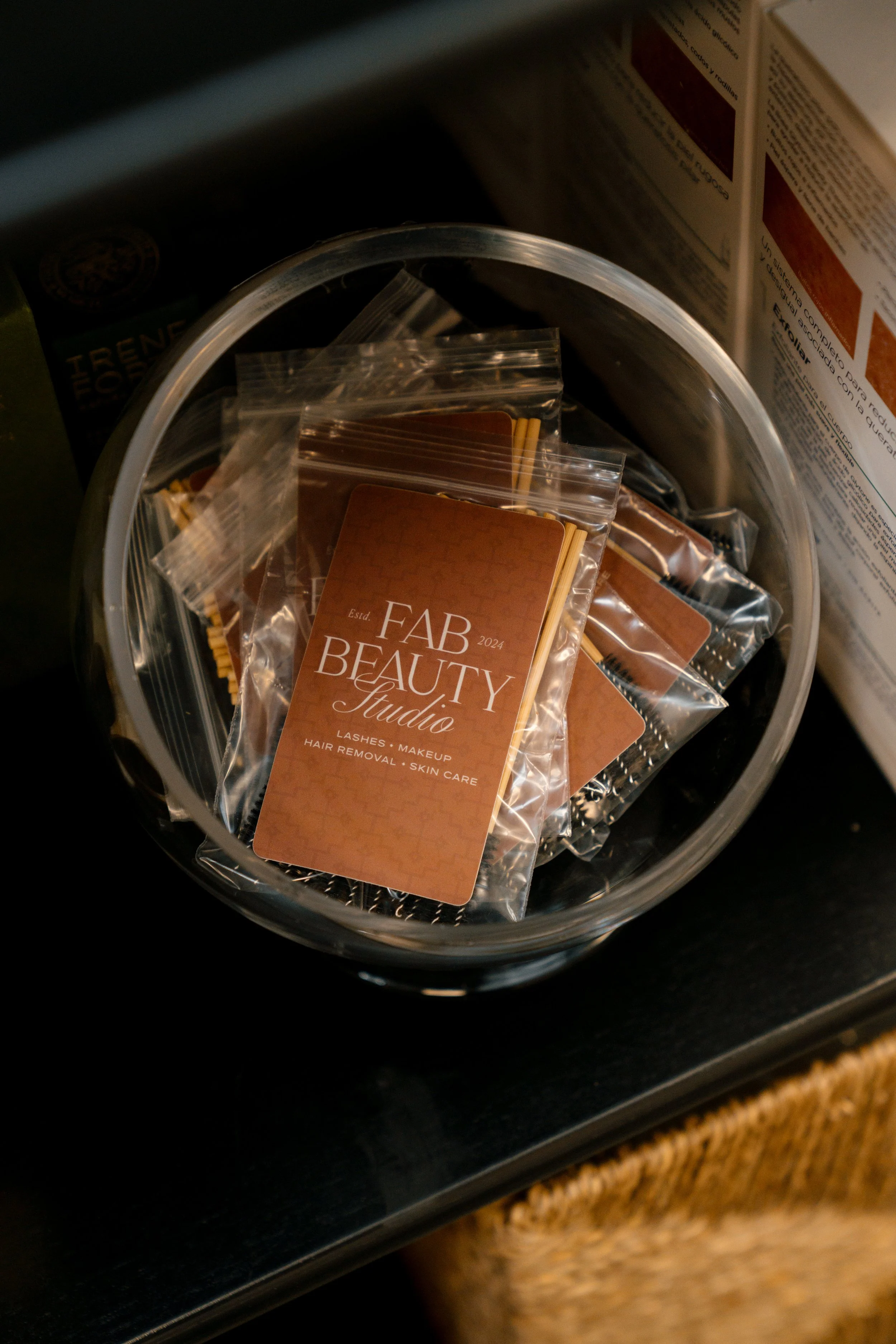 A clear round container holding small packets labeled 'FAB Beauty Studio' with services like lashes, makeup, hair removal, and skin care written on the packets.