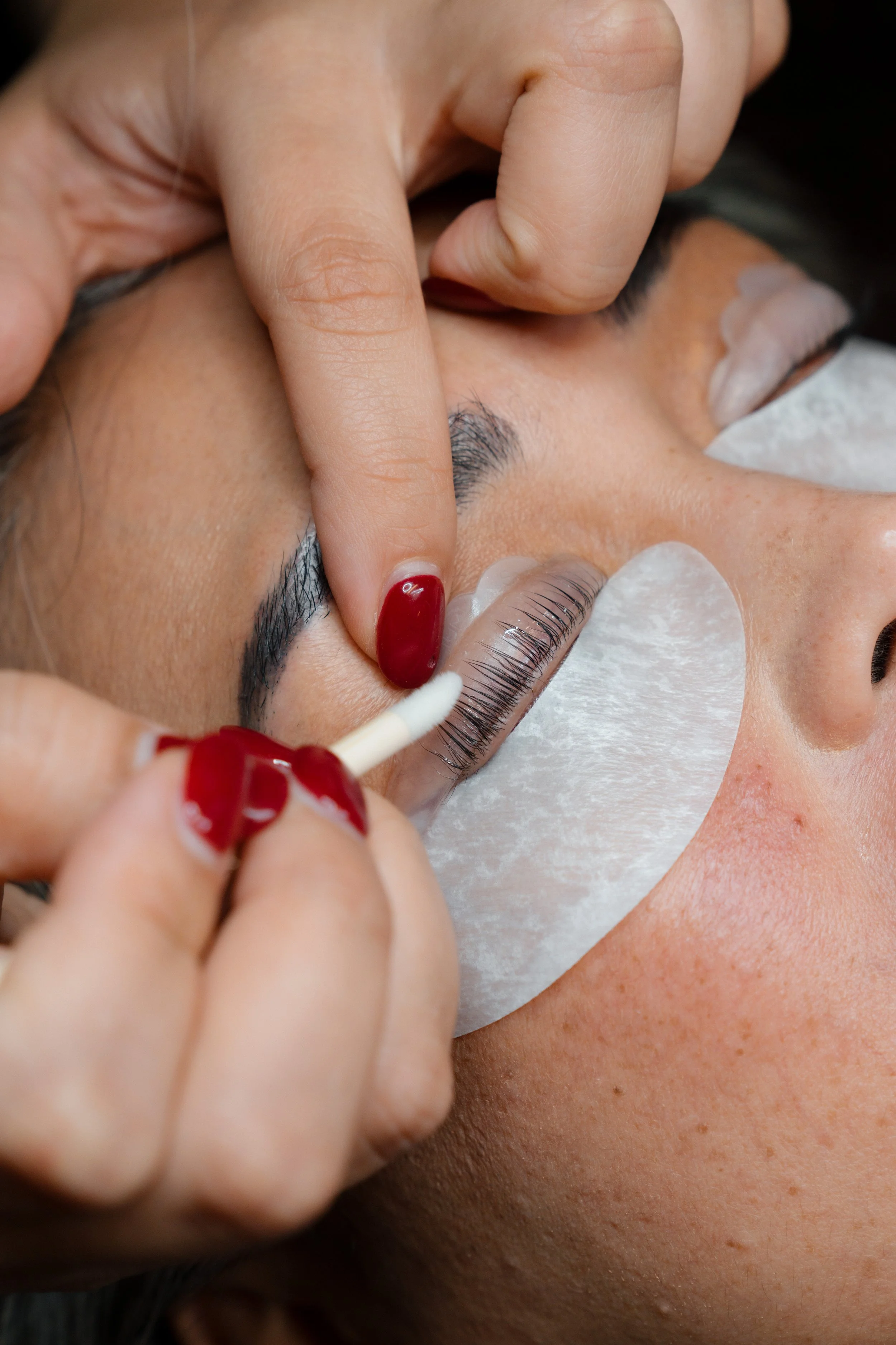 Close-up of a person receiving eyelash extension application, with a technician using a cotton swab and adhesive, under eye patches.