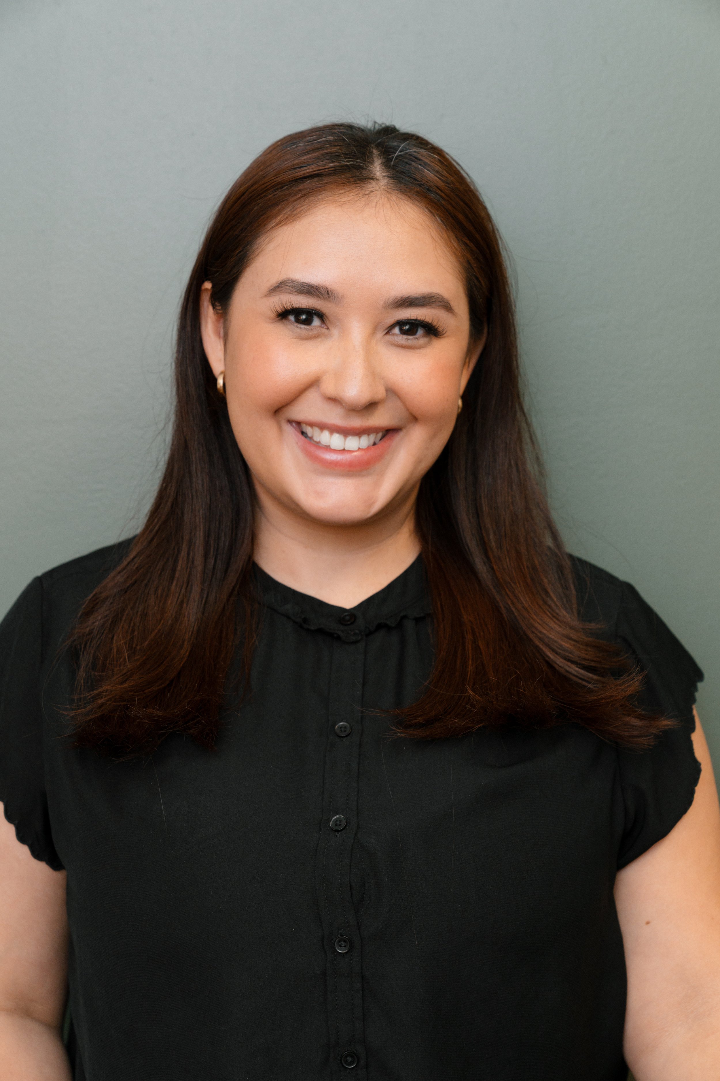 A woman with shoulder-length brown hair and a black blouse smiling at the camera against a grey background.