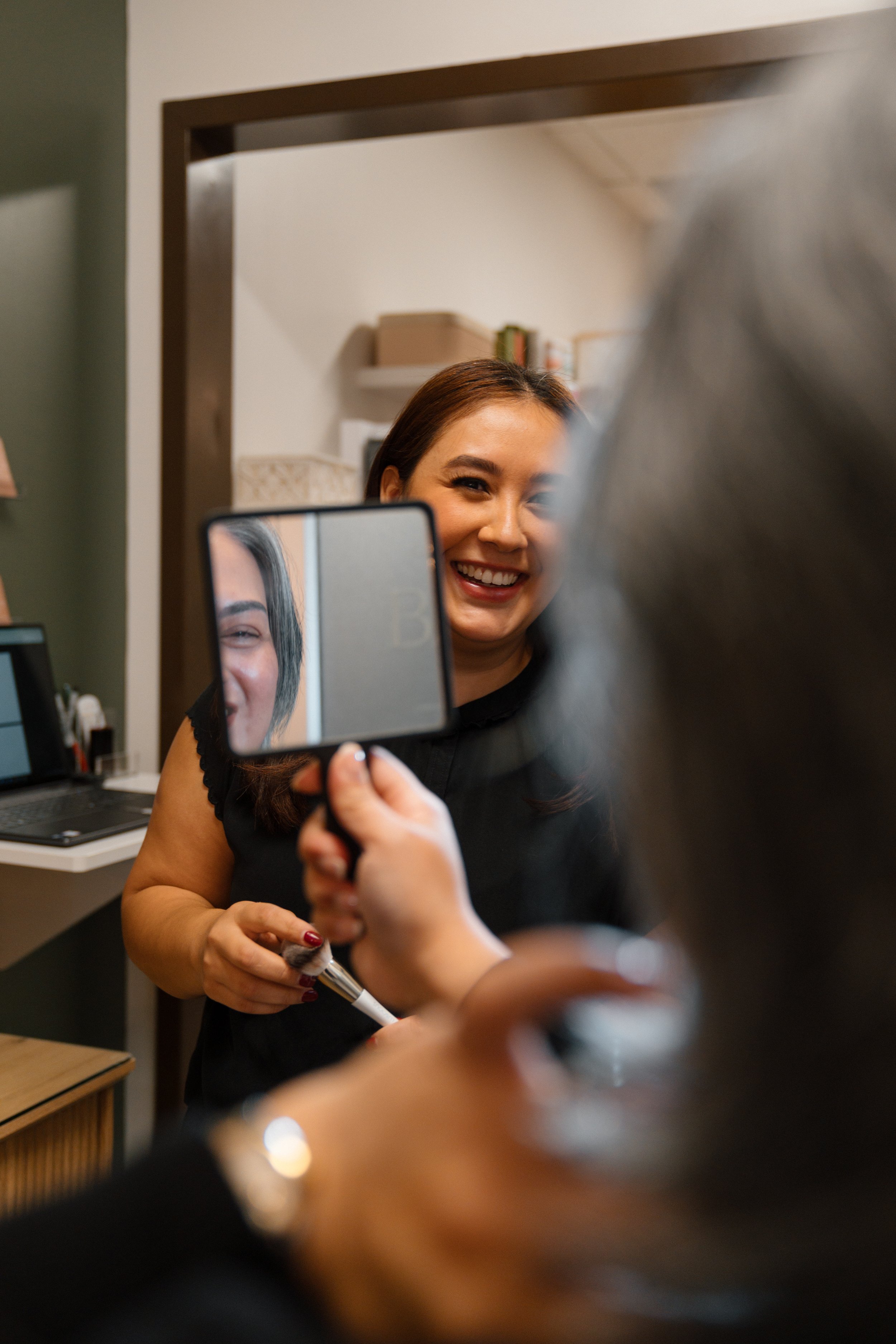 A woman smiling at her reflection in a mirror while another person takes a photo with a handheld mirror.