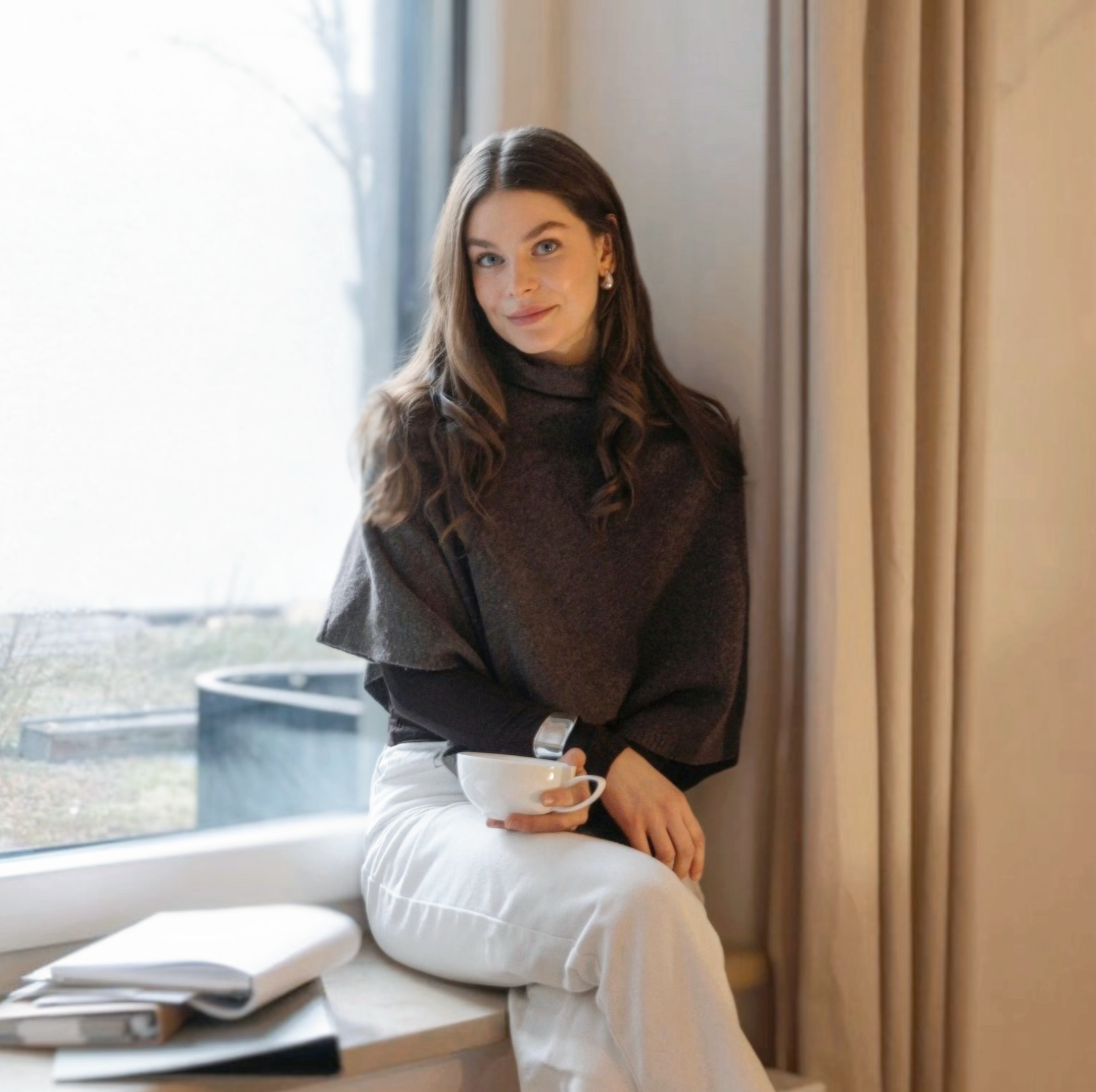 Junge Frau mit braunen Haaren sitzt am Fenster, hält eine Tasse und schaut lächelnd in die Kamera, im Hintergrund Gardinen.