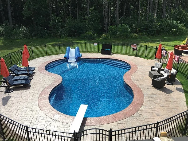 Backyard with a kidney-shaped swimming pool, slide, patio furniture, umbrellas, and a surrounding black metal fence, with trees in the background.