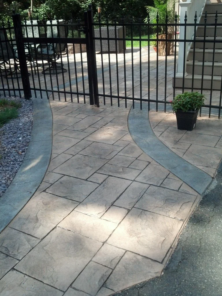 Black metal fence gate on a stone paved patio with a potted plant and outdoor furniture, leading to a grassy backyard area.