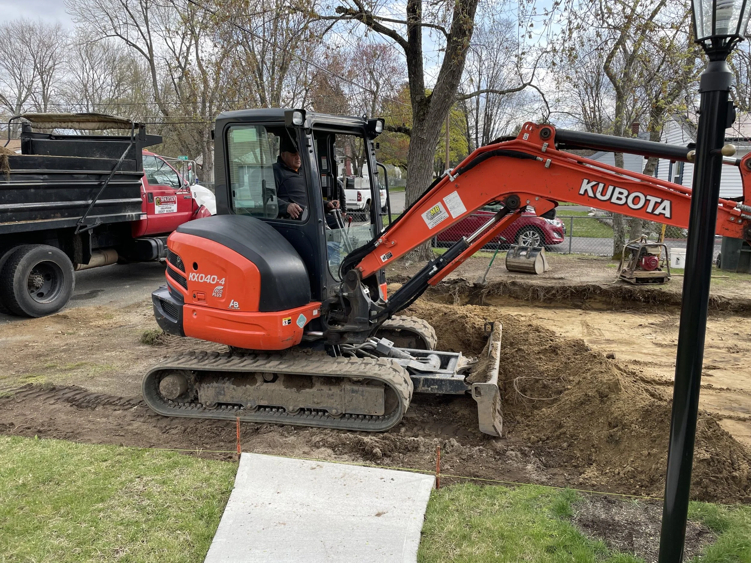 A person operating a Kubota mini excavator digging a trench in a residential backyard with a sidewalk at the entrance and street with parked vehicles in the background.