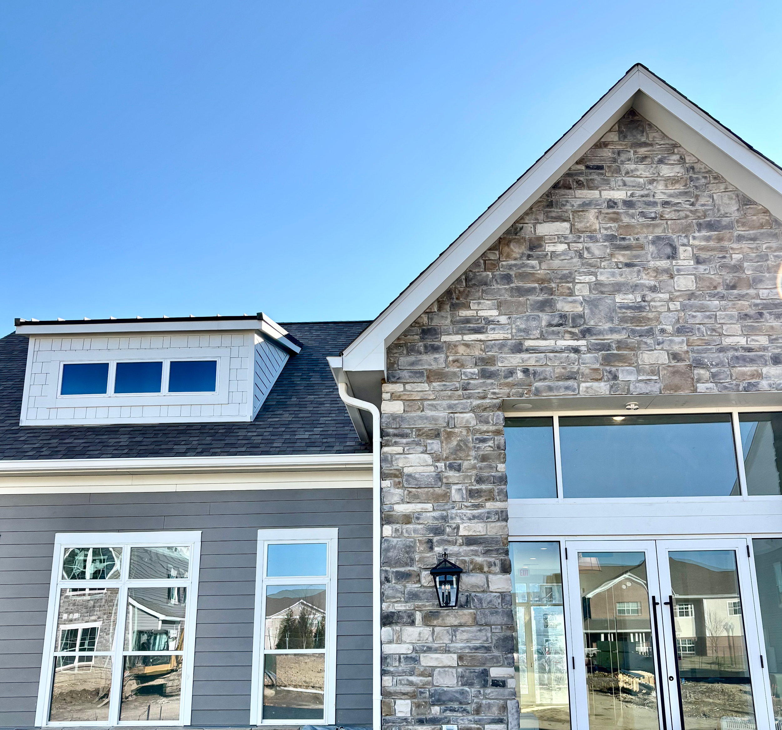 Close-up view of Carriage Park Apartment's clubhouse house with a stone and gray siding exterior, large windows, and a gabled roof under a clear blue sky.