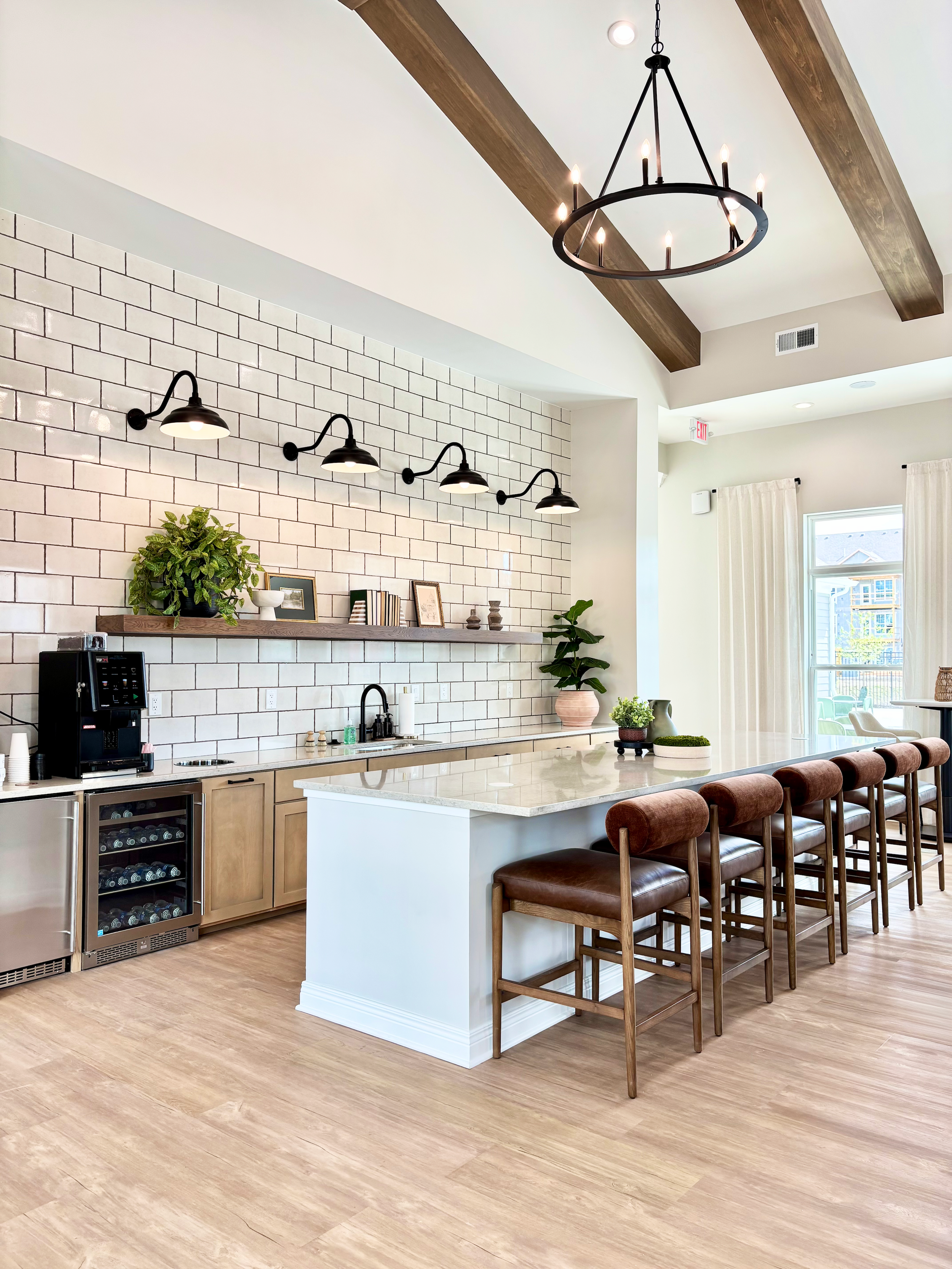 Modern kitchen with white cabinets, beige countertop, white tile backsplash, bar area with brown stools, plants, and black hanging light fixture.