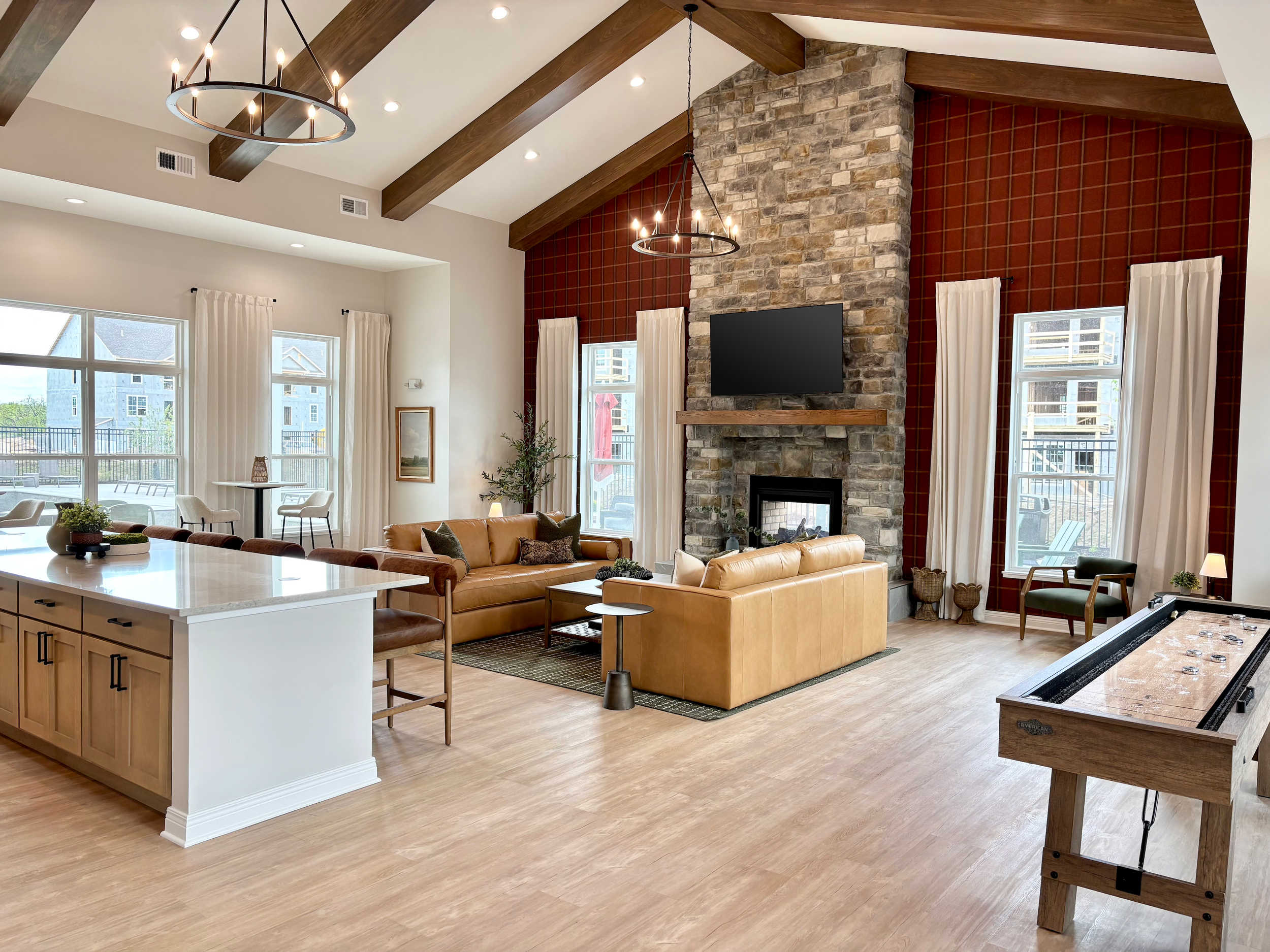 Open-concept living room with wood-beamed ceiling, stone fireplace with mounted TV, tan leather sofas, and large windows with white curtains.