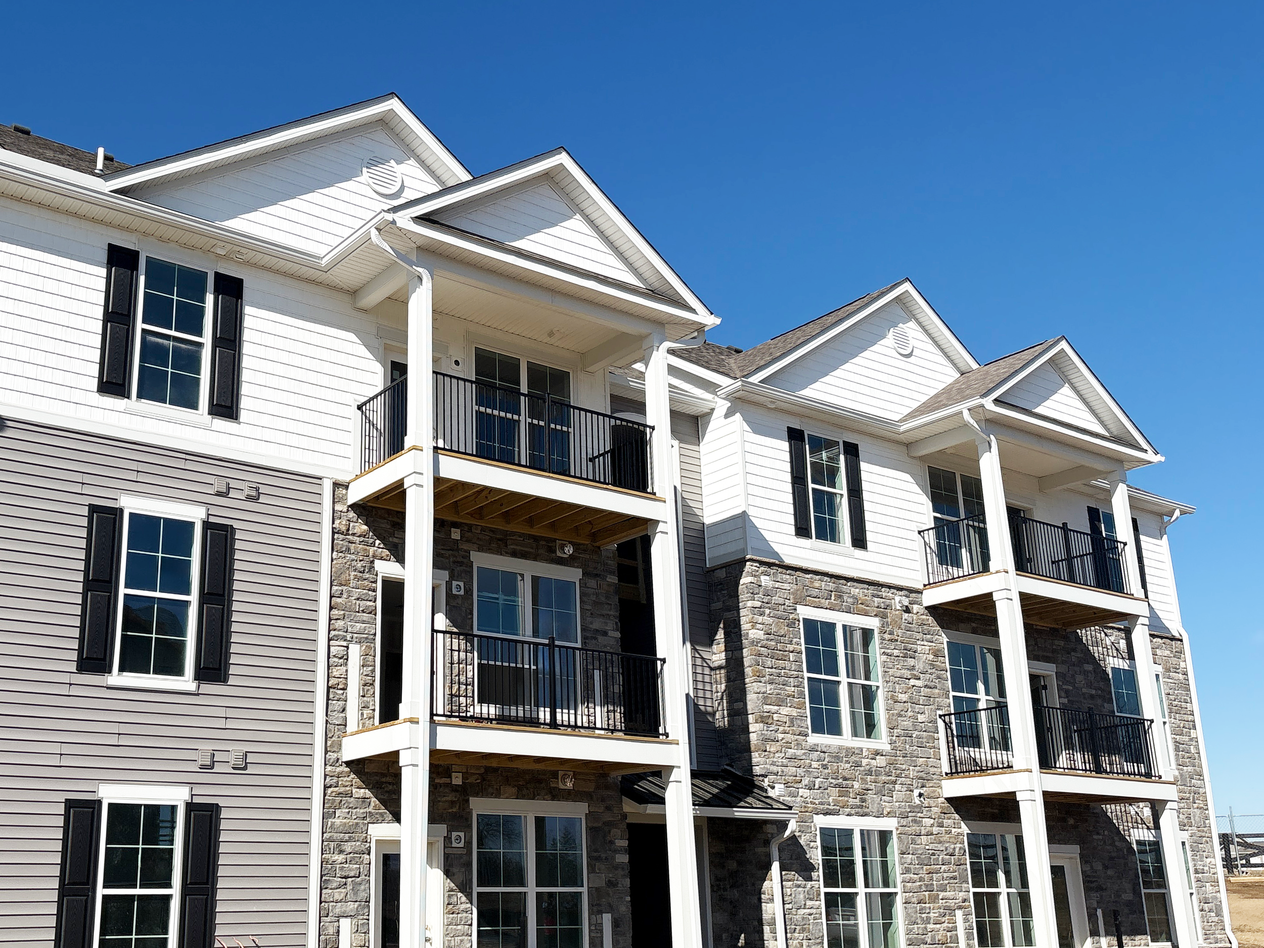 A three-story multifamily apartment building with white siding and stone accents, featuring balconies with black railings and large windows, under a clear blue sky.