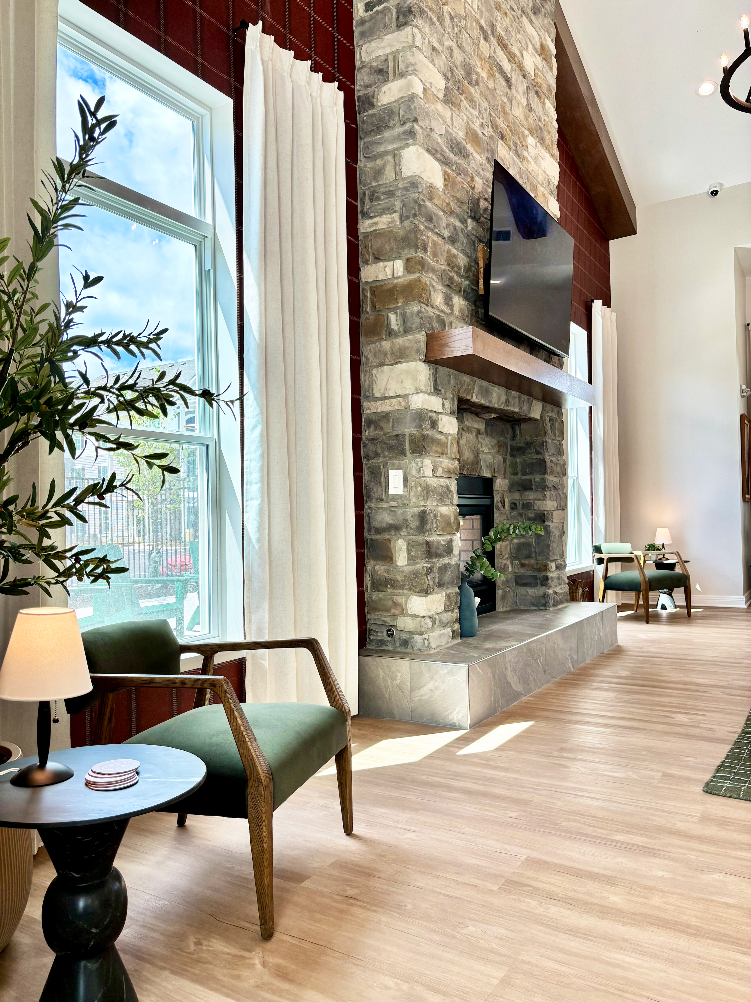 Living room with a stone fireplace, flat-screen TV above the mantel, green chairs, a small table with a lamp, and large windows with white curtains allowing sunlight in.