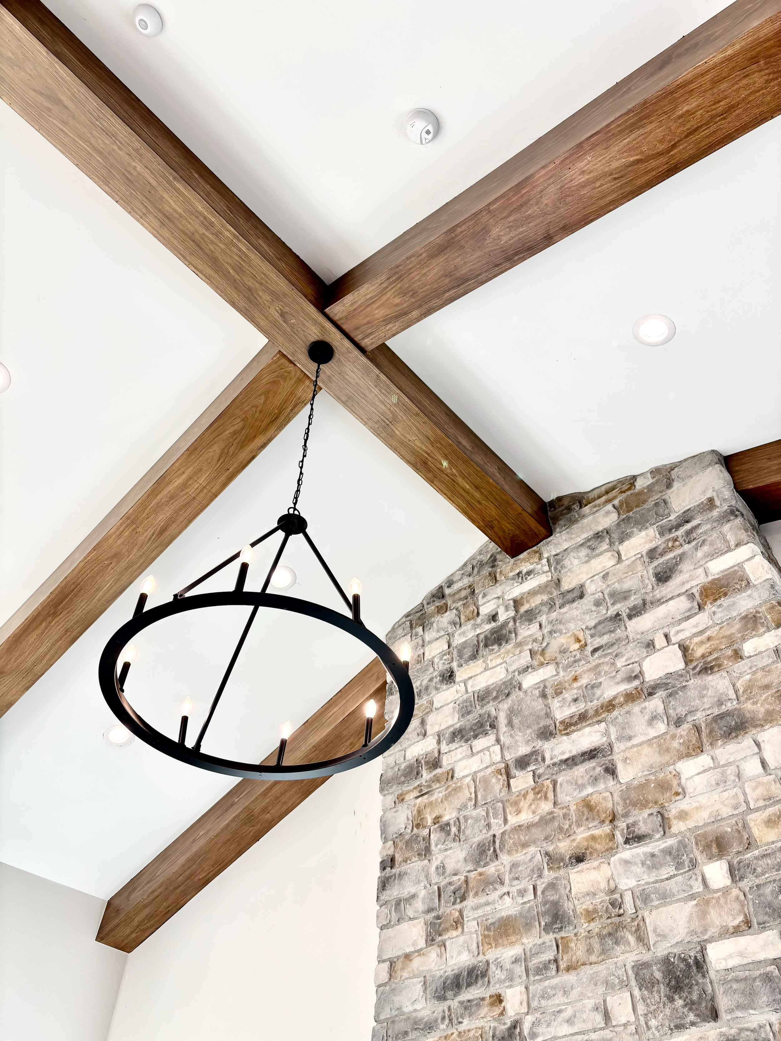 Interior ceiling view featuring exposed wooden beams, a hanging black circular chandelier with candle-like light fixtures, and a brick chimney or wall segment.