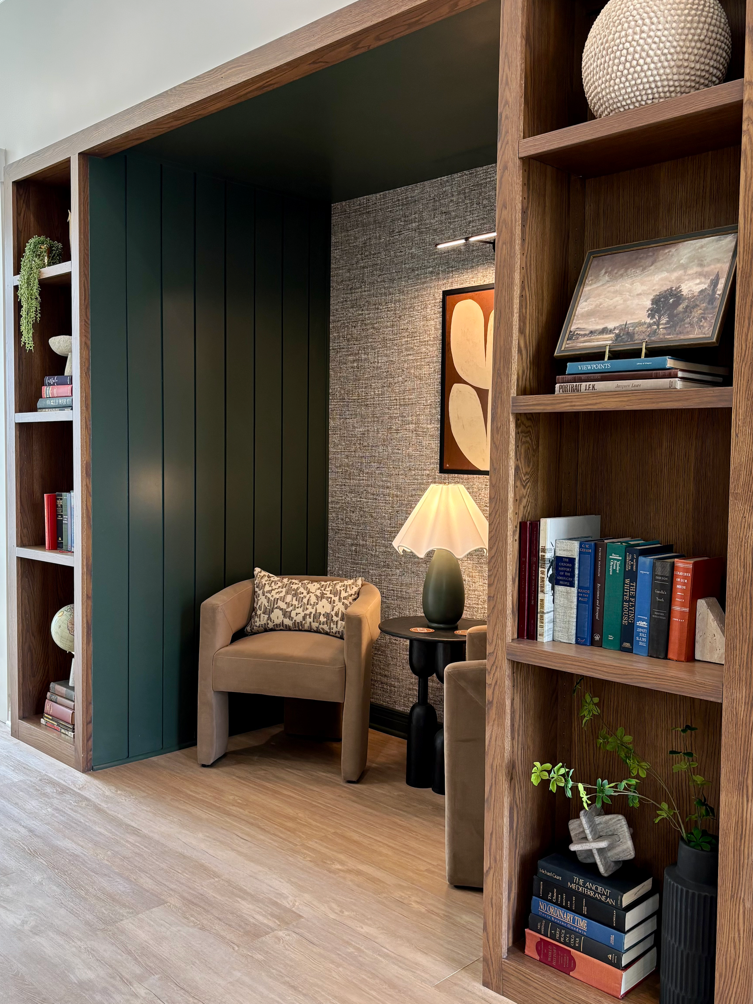 A cozy living room corner with a beige armchair, a lamp on a small black side table, and built-in wooden shelves filled with books and decorative items. There is a textured wall with artwork hanging behind the armchair and a light-colored wood floor.