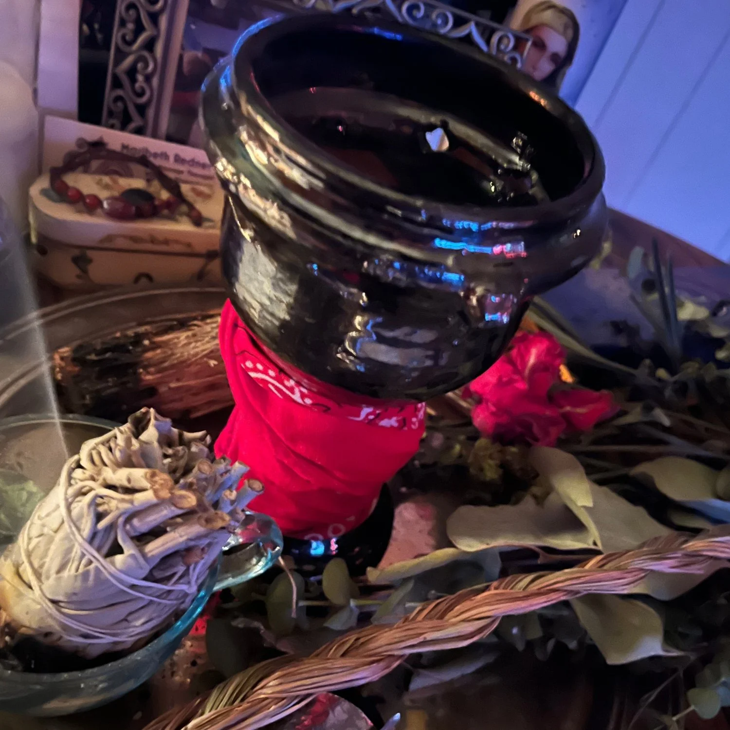 A person's arm and hand holding a large, empty, black chalice wrapped in a red cloth. The scene includes floral arrangements, dried mushrooms, a bottle, and a framed picture of a woman in the background.