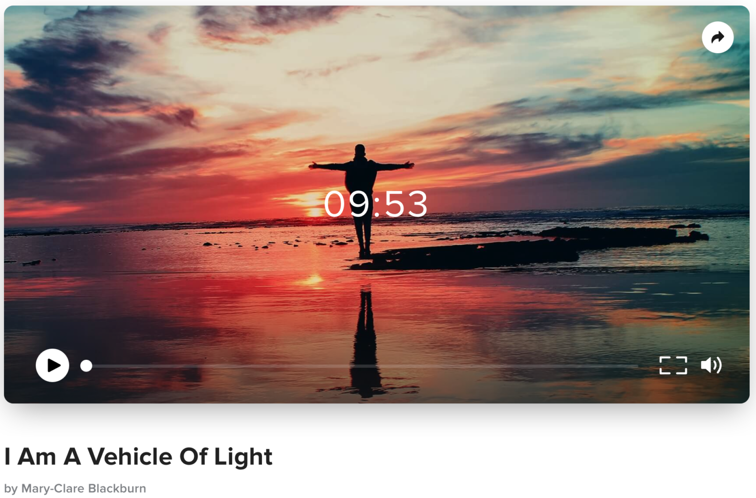 A person standing on a rocky beach with arms outstretched during sunset, with vibrant orange, pink, and blue colors in the sky, and the person's reflection visible in the shallow water.