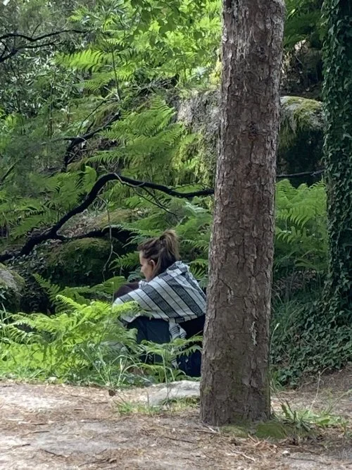 A woman sitting on the ground in a lush green forest, partially obscured by a tree in the foreground.