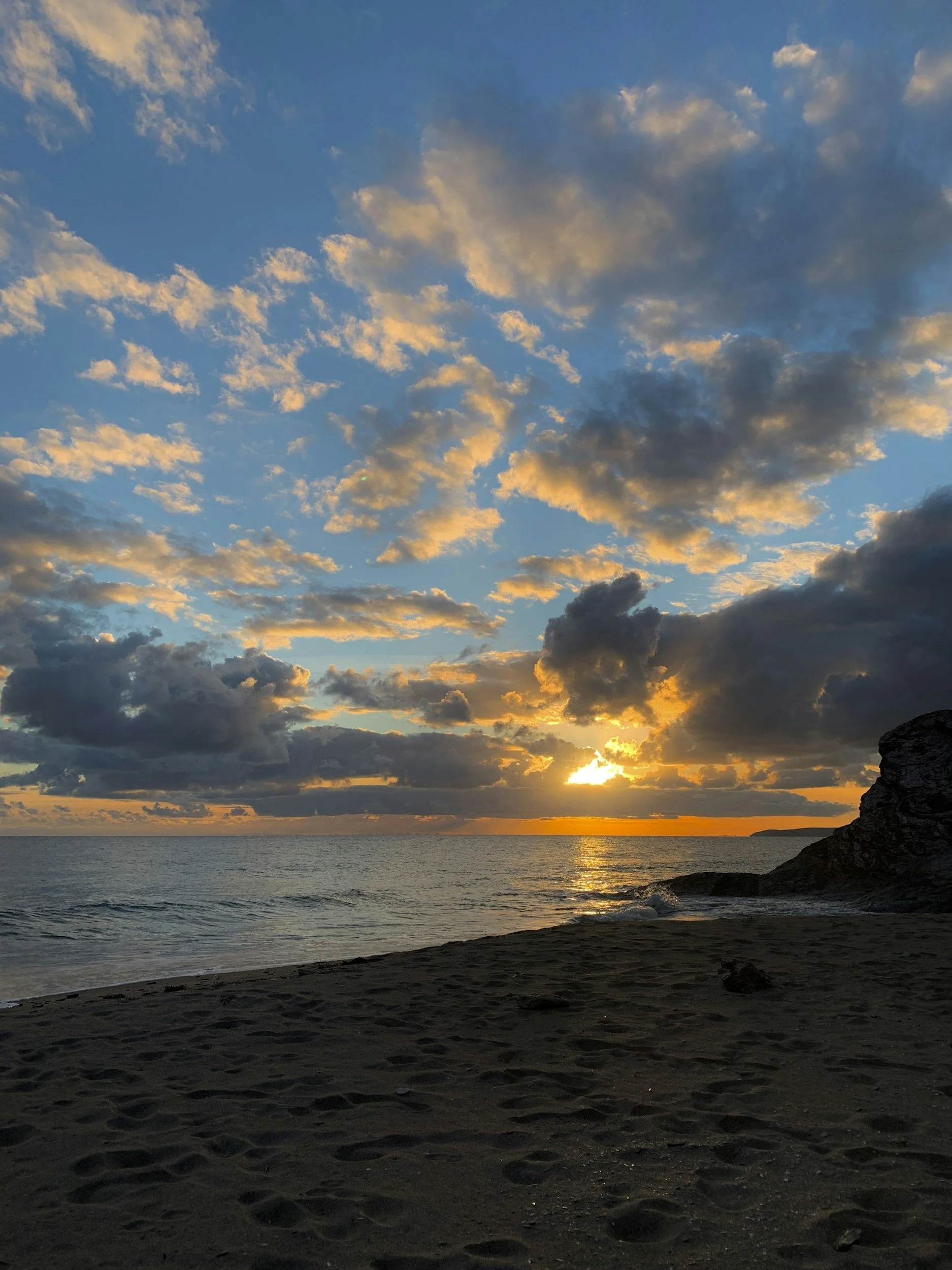 Sunset over the ocean with partly cloudy sky and a rocky shoreline.