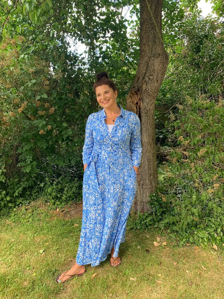 Woman in a blue patterned maxi dress standing outdoors near a tree, smiling.