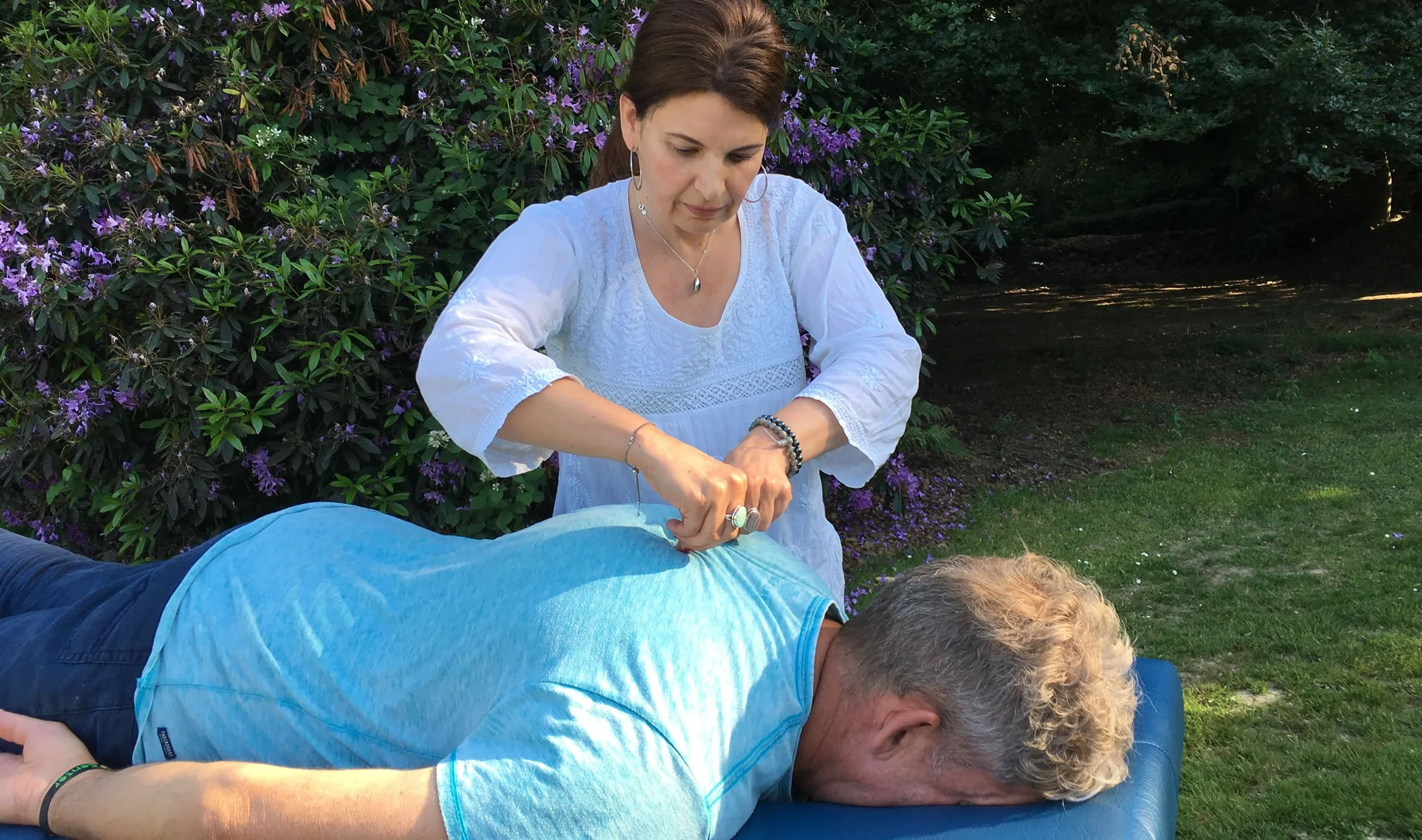 A woman giving an outdoor massage to a man lying face down on a massage table, with purple flowers and green bushes in the background.