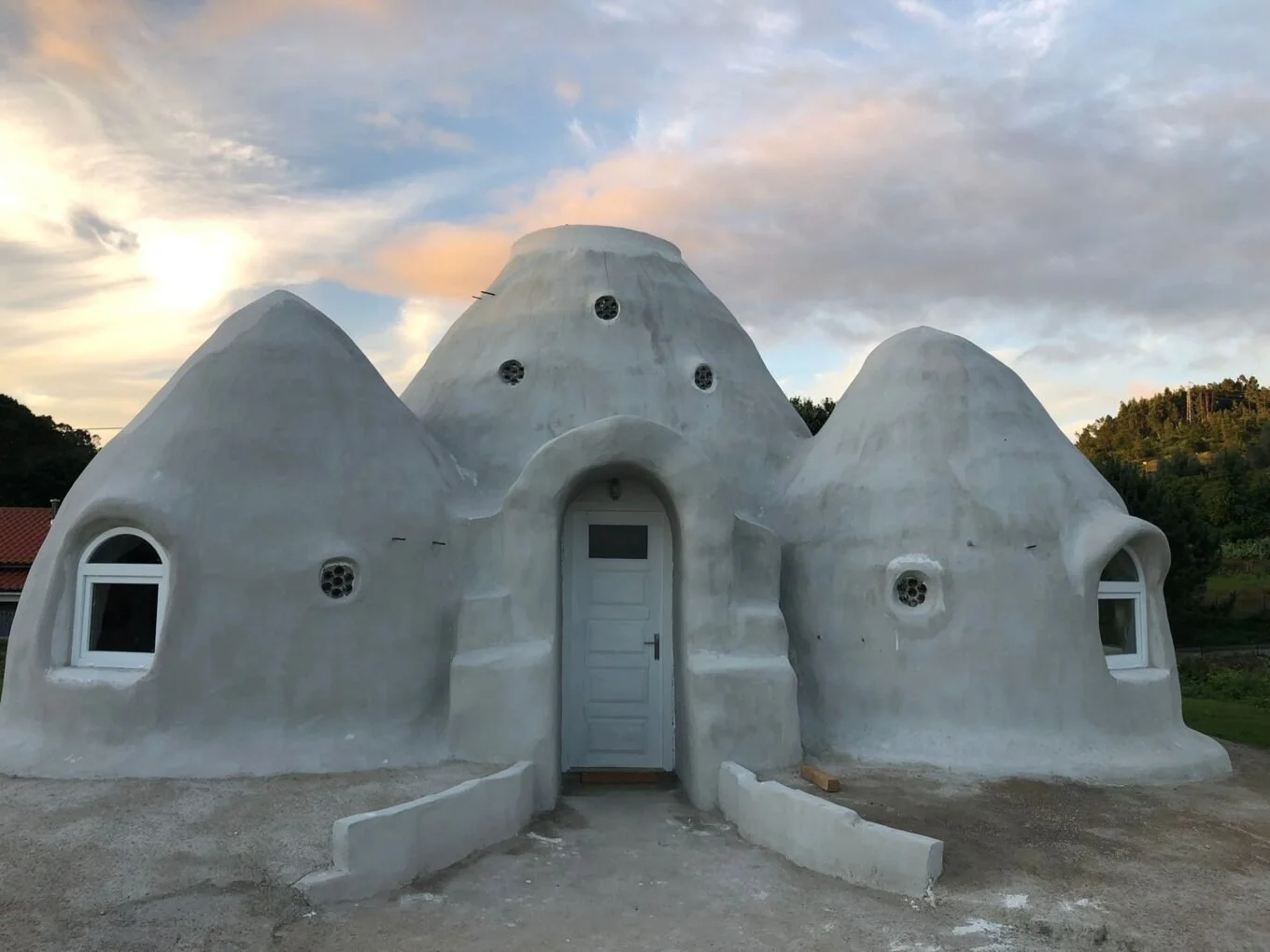 White, organic-shaped house with rounded, irregular walls and small circular and arched windows, set against a sunset sky with clouds and trees in the background in Portugal.