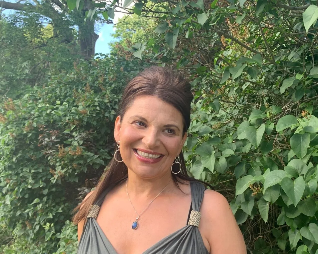 A woman smiling outdoors in front of green foliage, wearing a gray dress, earrings, and a necklace with a blue pendant.
