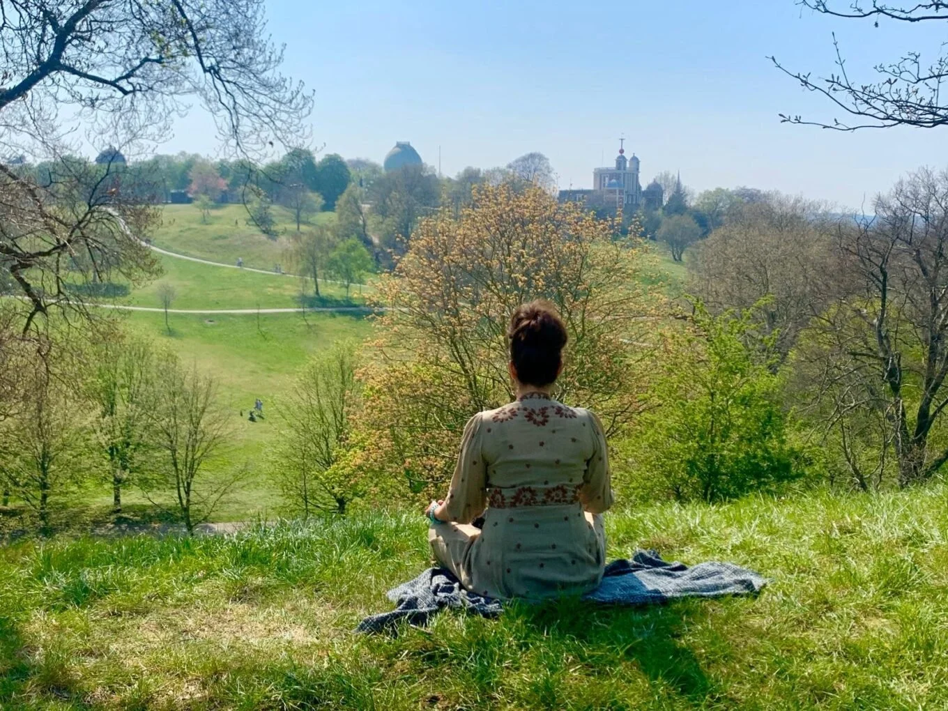 A woman sitting on a grassy hill overlooking a landscape with trees, pathways, and buildings in the distance on a sunny day.