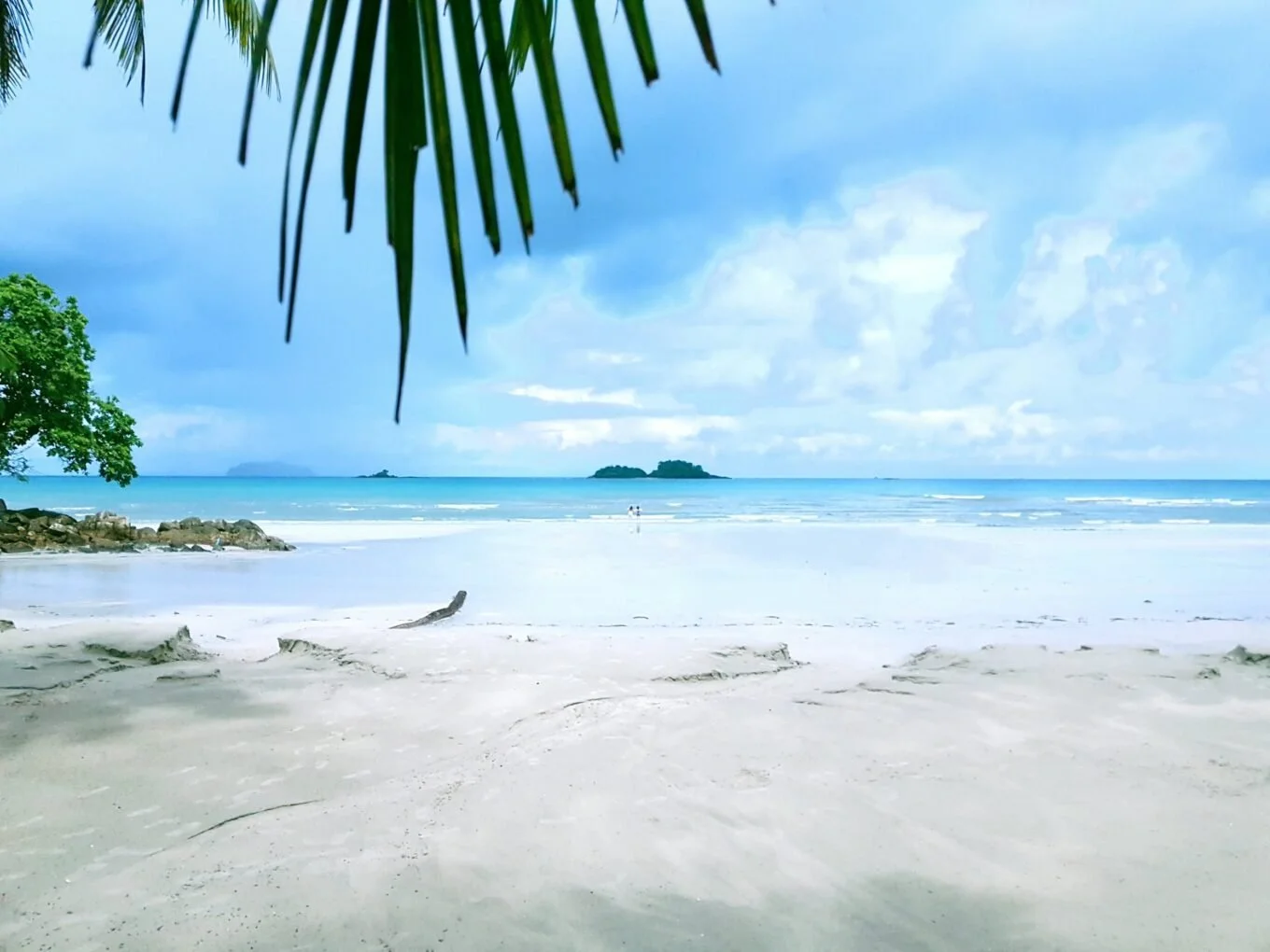 Tropical beach with white sand, calm ocean, green trees, and an island in the distance under a partly cloudy sky.