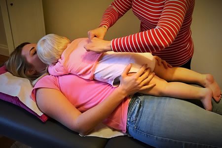 A woman lying on her back on a massage table, with a young child lying across her chest. An adult standing beside them appears to be giving a massage to the child.