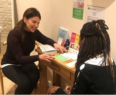 A woman with long dark hair talking to another woman with dreadlocks at a small table inside a room.