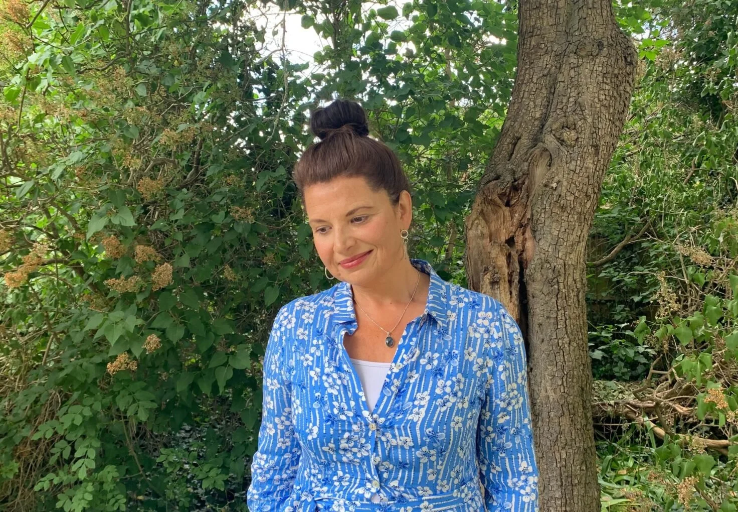 A woman with brown hair tied in a bun, wearing a blue floral shirt with a white undershirt, standing next to a tree in a lush green environment, smiling softly and looking down.