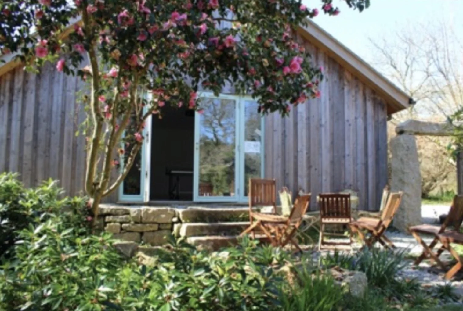 A wooden house with an open door and large window, surrounded by greenery and pink flowering trees, with outdoor wooden chairs and tables on a stone patio in Cornwall.