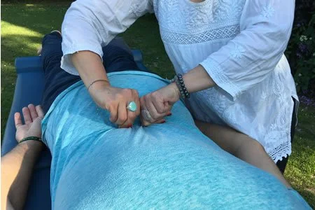 Person receiving a massage outdoors on a massage table, with greenery nearby, while another person performs the massage.