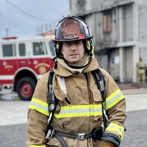 Firefighter in gear standing in front of a fire truck and a building.