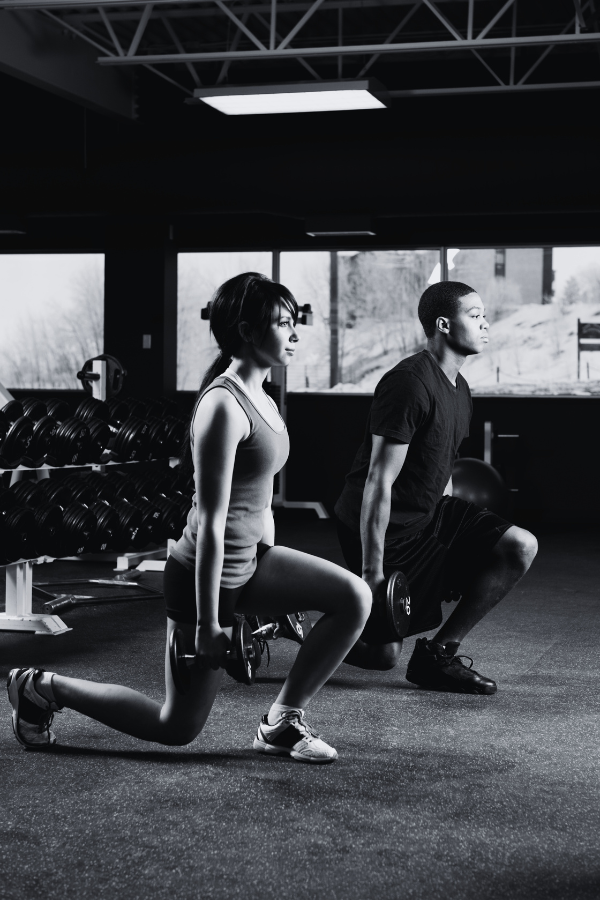 Two people exercising in a gym with dumbbells, performing lunges while holding weights, in front of large windows with outdoor scenery.