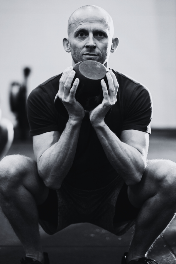 A man with a shaved head and athletic build in workout attire holding a kettlebell in a gym.