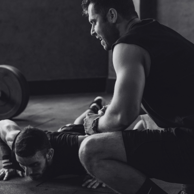 A man assisting a woman with a workout in a gym, both on the floor, with gym equipment in the background.