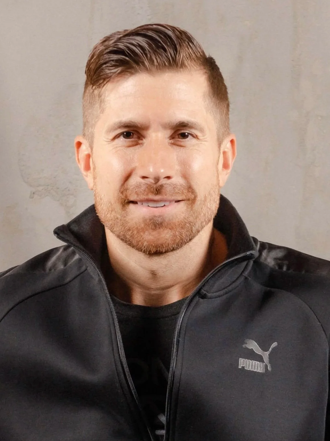 A man with short brown hair and a beard, wearing a black Puma jacket, smiling and looking at the camera.