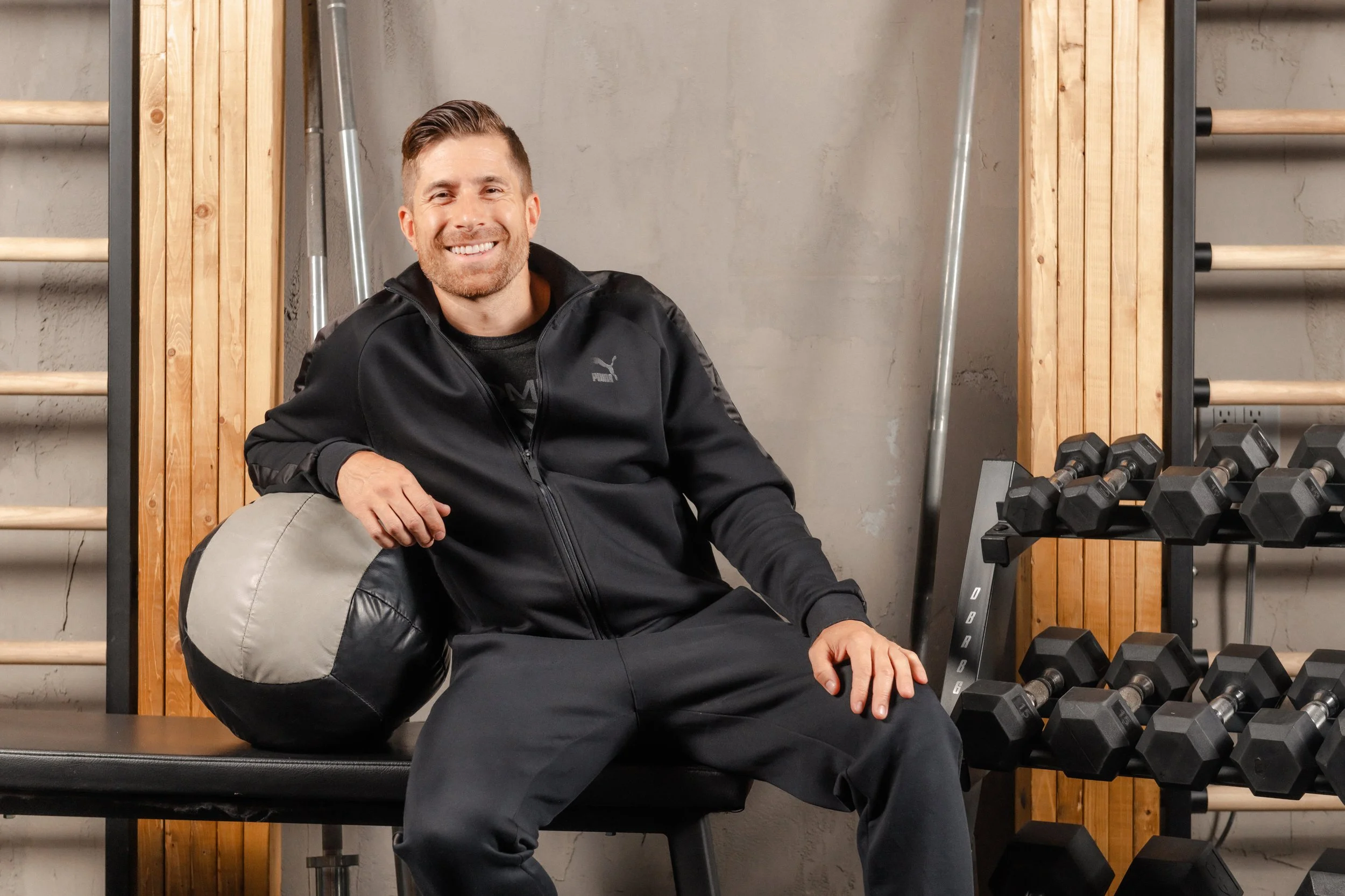 A smiling man sitting on a workout bench in a gym, holding a gray and black medicine ball, with a set of dumbbells and wooden wall bars in the background.