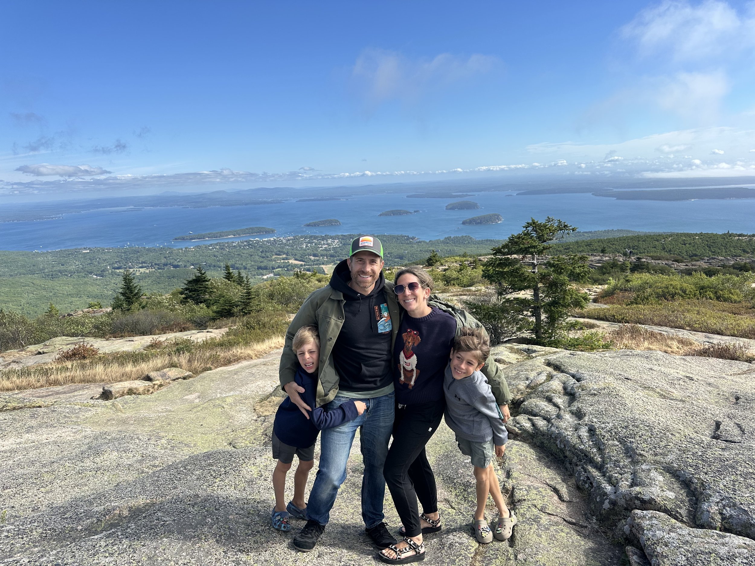 A family of four hiking on a rocky trail with a scenic view of a lake and islands, under a partly cloudy sky.