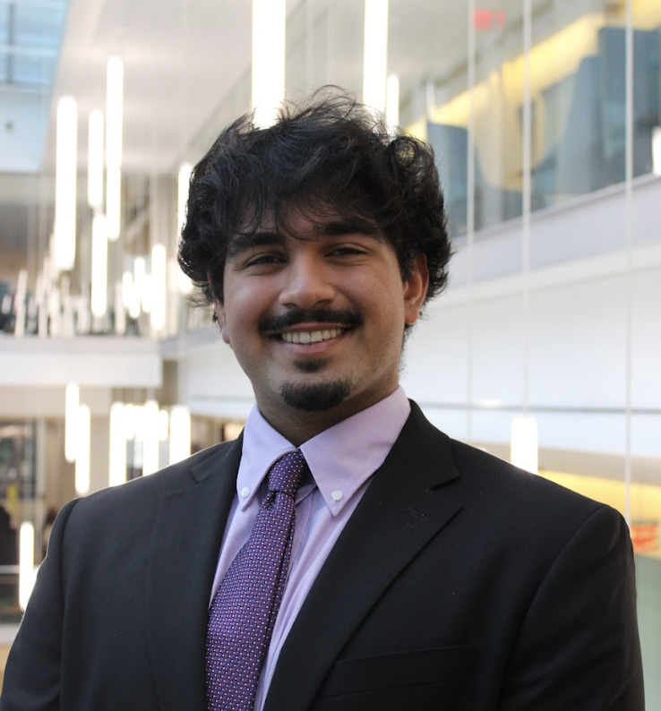 A smiling man in a black suit and purple tie standing inside a modern building with large glass windows and interior lighting.