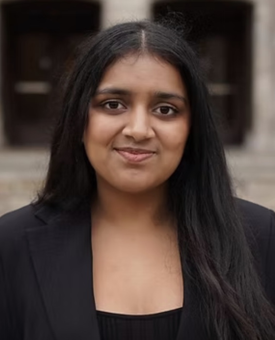 A woman with long black hair, wearing a black blazer and top, standing outdoors in front of a building with stone steps.
