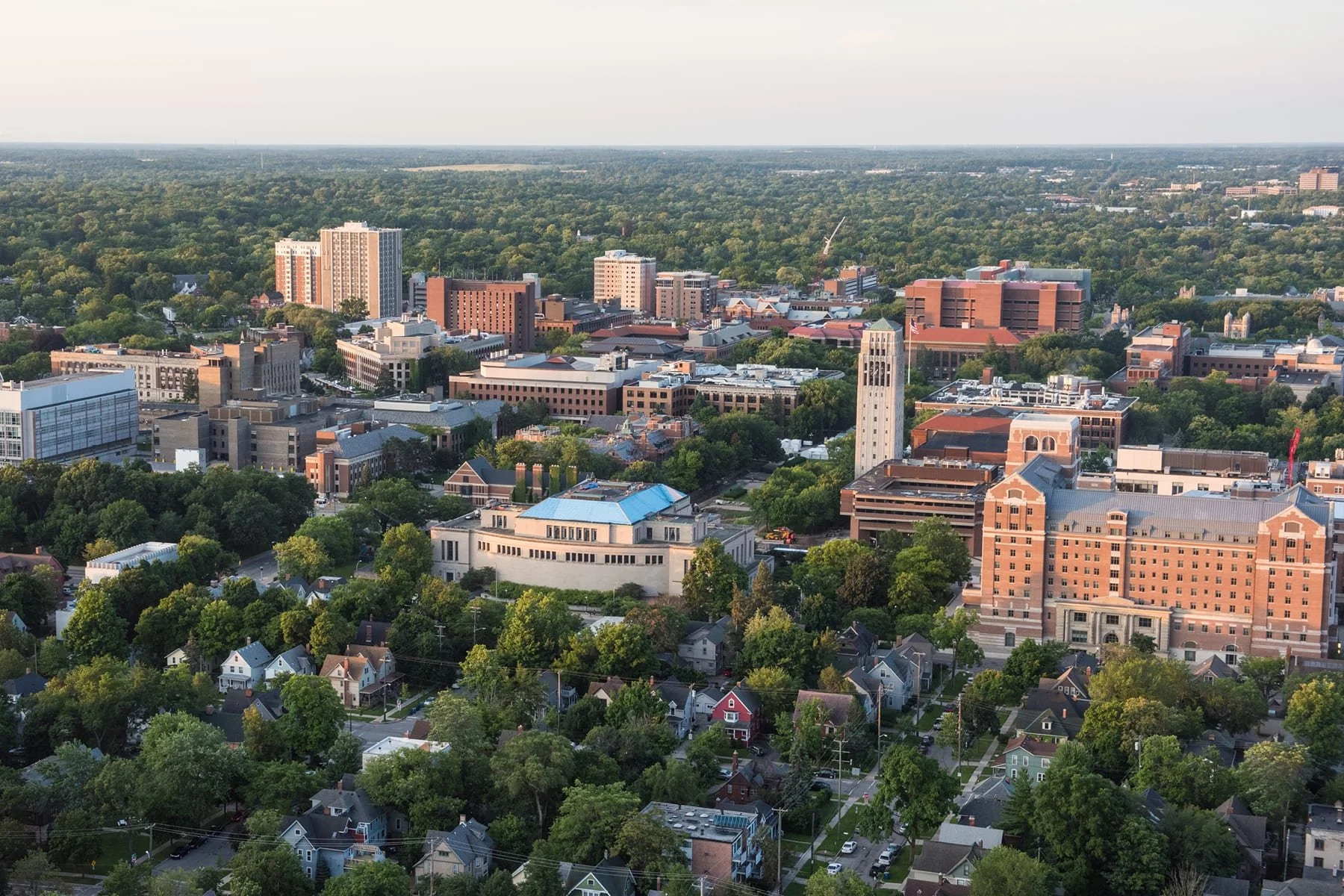Aerial view of a cityscape with a mix of residential houses, trees, and taller office buildings and a clock tower, with a wooded area extending to the horizon.