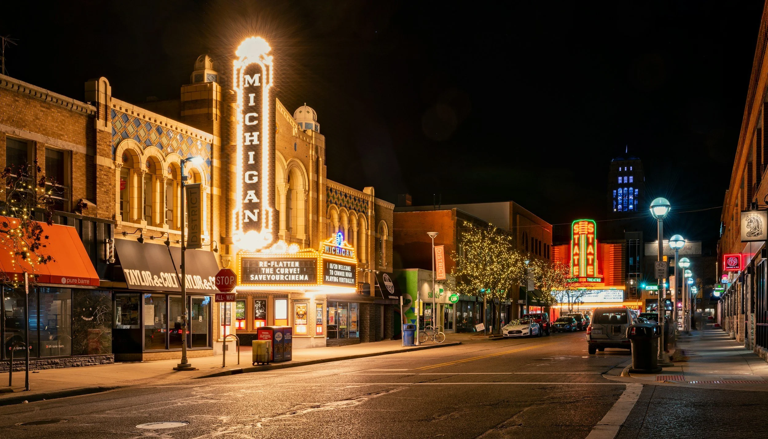 Night scene of a downtown street with illuminated neon theater signs, storefronts, Christmas tree lights, parked cars, and street lamps.