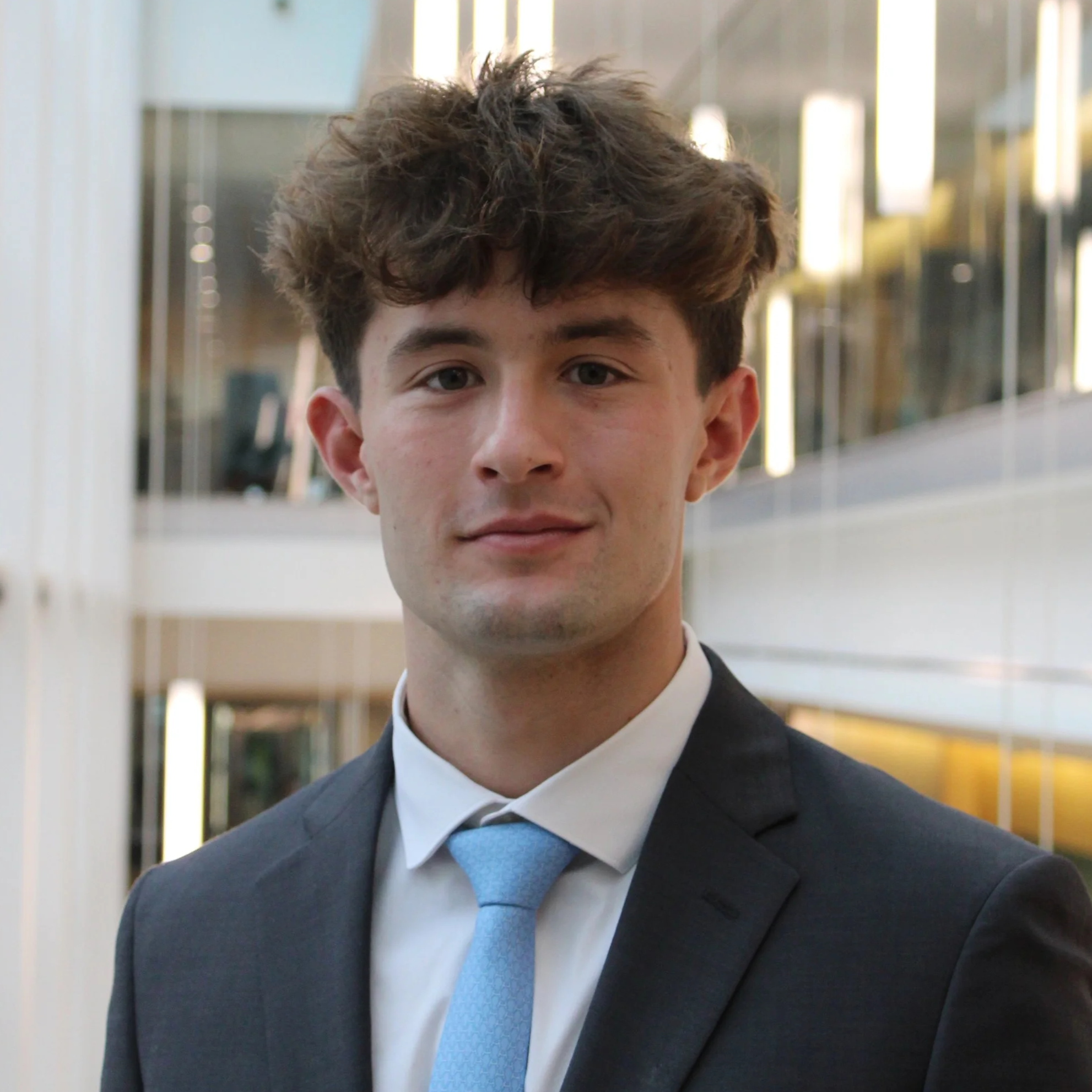 A young man in a dark suit and light blue tie standing indoors in a modern building.