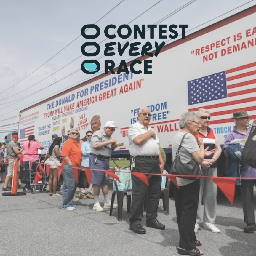 People standing in line at an outdoor event with a large political campaign billboard in the background.